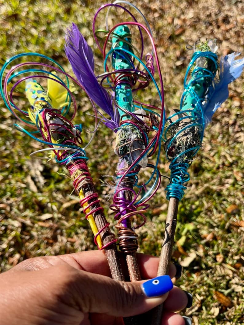 May include: Three wooden wands wrapped in colorful wire, feathers, and crystals. The wands are held in a hand with blue nail polish.