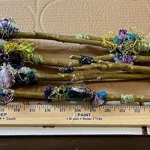 May include: A collection of 10 thin wooden sticks wrapped in colorful fabric scraps and yarn. The sticks are arranged on a brown surface with a ruler measuring 27 inches.