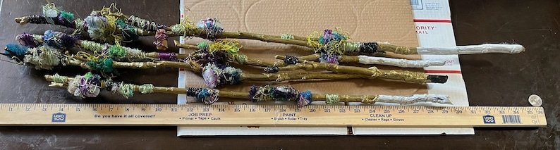 May include: A collection of 10 thin wooden sticks wrapped in colorful fabric scraps and yarn. The sticks are arranged on a brown surface with a ruler measuring 27 inches.