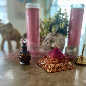 May include: A pink and gold resin pyramid with a small, ornate, dark brown bottle with colourful gems on a table with two pink candles and a gold bell.