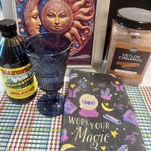 Könnte beinhalten: Eine Auswahl an Artikeln, darunter ein dunkelblaues Glas, eine Flasche Plantation Blackstrap Melasse, ein Glas Ceylon-Zimt und ein Notizbuch mit dem Text "WORK YOUR Magic". Im Hintergrund befindet sich ein gerahmtes Bild von Sonne und Mond.