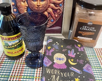 May include: An assortment of items including a dark blue goblet, a bottle of Plantation Blackstrap Molasses, a jar of Ceylon Cinnamon, and a journal with the text "WORK YOUR Magic". A framed picture of the sun and moon is in the background.