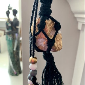 May include: A black macrame hanging with a gold and white painted rock, black tassels, and wooden beads in gold, white, and gray.