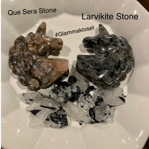 Peut inclure: Deux pierres en forme de licorne, l'une à motif brun clair et blanc étiquetée "Que Sera Stone" et l'autre à motif gris foncé étiquetée "Larvikite Stone". Les pierres sont entourées de petites pierres blanches et noires. Le hashtag "#Glammakloset" est écrit sous les pierres.