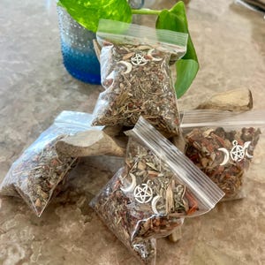 May include: Four clear plastic bags filled with dried herbs and small silver charms. The charms are pentacles and crescent moons.