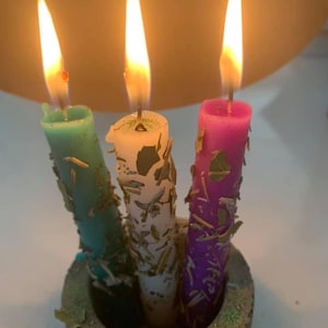 May include: Three candles in a concrete holder. The candles are teal, white, and purple. Each candle has gold flakes embedded in the wax. The candles are lit.