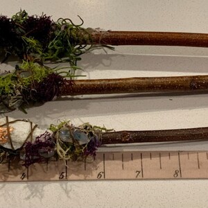 May include: Three wooden wands wrapped with moss, crystals, and other natural elements. The wands are approximately 7 inches long and are displayed on a ruler.