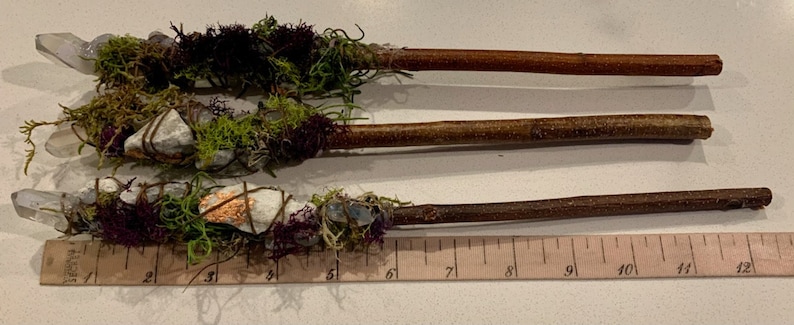 May include: Three wooden wands wrapped with moss, crystals, and other natural elements. The wands are approximately 7 inches long and are displayed on a ruler.