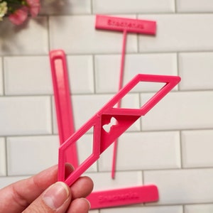 Könnte beinhalten: Eine Hand hält ein leuchtend rosa geometrisches Werkzeug mit einem ausgeschnittenen Bogen. Das Werkzeug ist Teil eines Sets, mit anderen sichtbaren Teilen, darunter ein langer, dünner Pfahl und ein rechteckiges Stück mit dem Text "Shachenka". Der Hintergrund ist eine weiße Fliese.