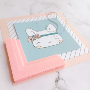 Könnte beinhalten: Ein hellrosa, eckförmiges Bastelwerkzeug mit dem Text "EASY SCRAP FOR YOU" liegt auf einer hellblauen Karte mit einem Cartoon-Hasen mit floralem Stirnband. Die Karte ist auf gestreiftem und unifarbenem Papier geschichtet.