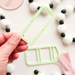 Può includere: Un tagliabiscotti rettangolare verde chiaro con piccoli dettagli decorativi è tenuto in mano. Un altro tagliabiscotti simile è sotto. Ghirlande di pompon bianchi e perline nere sullo sfondo.