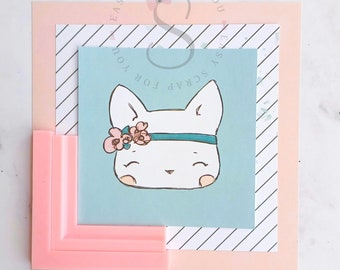 May include: A cute illustration of a white cat face with a floral headband and a teal band. The cat is on a light blue background, layered with a striped paper and a pink frame. The image is a square composition.