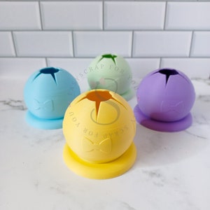 May include: Three silicone molds in the shape of a sphere with a bow design. The molds are light blue, yellow, and purple.