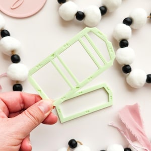 Puede incluir: Dos plantillas rectangulares de manualidades en forma de etiqueta de color verde claro se sostienen en una mano. Las plantillas tienen un diseño similar a una ventana con pequeños elementos decorativos. Guirnaldas de pompones blancos y cuentas negras en el fondo.