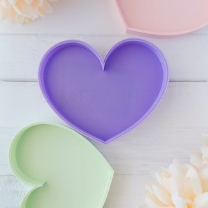 Puede incluir: Dos bandejas en forma de corazón, una verde claro y otra morada, sobre una superficie de madera blanca.