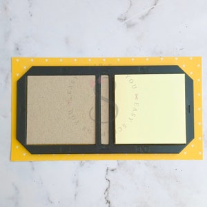 Op de afbeelding: Een zwarte plastic bureau-organizer met twee vakken. Het linker vak heeft een bruin papieren notitieblok en het rechter vak heeft een geel kleefnotitieblok. De organizer staat op een gele achtergrond met een wit hartpatroon.