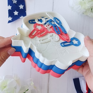 May include: A set of three red, white, and blue plastic cookie cutters shaped like a star. The cutters are stacked on top of each other and are surrounded by red, white, and blue paper clips.