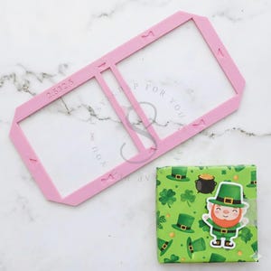 Può includere: Un modello rosa per lavori di artigianato con il testo "2.532.5" e una confezione regalo a tema San Patrizio. La confezione regalo è avvolta in carta verde con trifogli, cappelli da folletto e un adesivo di folletto.