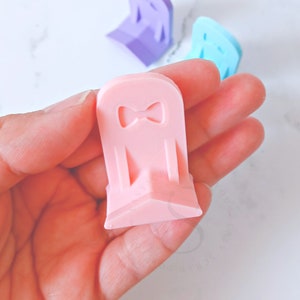 May include: A pink plastic chair with a bow on the back. The chair is small and has a triangular base.