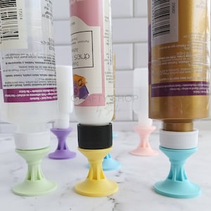 Peut inclure: Cinq tubes de peinture sont debout sur des bouchons de bouteille en silicone de couleur. Les bouchons ont une base large et un col étroit. Les tubes de peinture sont de différentes couleurs, dont l'or, le blanc et le rose.