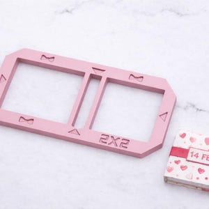Può includere: Una cornice rettangolare rosa chiaro con la scritta "2X2" e ritagli a forma di fiocco e triangolo. Accanto, un piccolo libro con un nastro rosso e la scritta "14 FEB" sulla copertina. Il libro è decorato con cuori.
