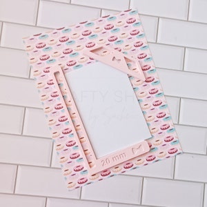 May include: Pink plastic frame with a cut-out for a 20 mm photo. The frame has a bow and heart design. The frame is on top of a pink and white patterned paper with donuts.