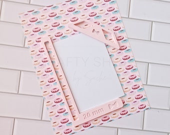 Pode incluir: Moldura de plástico rosa com um recorte para uma foto de 20 mm. A moldura tem um design de laço e coração. A moldura está sobre um papel estampado rosa e branco com donuts.