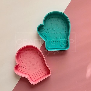 May include: Two plastic mitten-shaped cookie cutters. One is teal, the other is pink. Both have snowflake and line details. The cutters are on a pink and white background.
