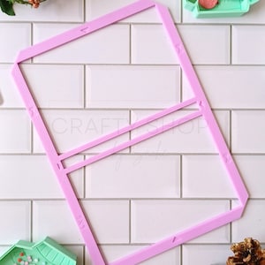 May include: A pink plastic template for creating a Christmas jumper design. The template has two horizontal lines and a vertical line. The template is on a white tiled surface.