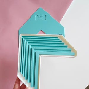 May include: A white plastic corner-shaped tray containing a stack of nested teal L-shaped plastic guides. Behind the tray is a flat teal template with a small bow-shaped cutout. The tray features multiple parallel grooves to hold the guides.