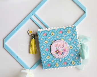 Puede incluir: Un pequeño cuaderno azul y blanco con estampado floral, ribete de pompón blanco y un círculo rosa con el texto "Wild at heart" en la parte delantera. El cuaderno tiene un borla amarilla y un amuleto de flor blanca. El cuaderno está sobre una superficie blanca con un marco de plástico azul y un contenedor de ojales de colores.