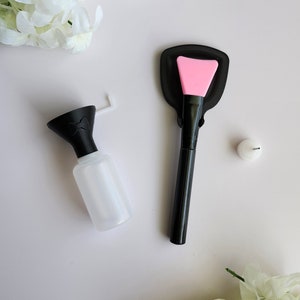 Könnte beinhalten: Draufsicht auf Beauty-Tools und weiße Blumen. Eine transparente Flasche mit schwarzem Deckel und ein rosa Applikatorpinsel sind zu sehen. Eine kleine weiße Kappe ist ebenfalls vorhanden. Die Artikel sind auf einer weißen Oberfläche angeordnet.