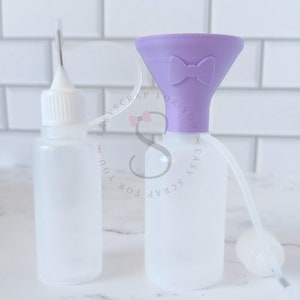 May include: Two clear plastic bottles with white caps. One bottle has a needle tip, the other has a purple funnel with a bow design. A clear tube connects the funnel to a bottle with a white cap. The bottles are for crafts.