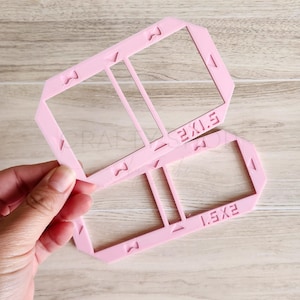 May include: A light pink plastic rectangular frame template with two open sections. The frame features embossed text reading 2X1.5 and 1.5X2, along with small triangular and M-shaped alignment markings along the edges of the frame structure.