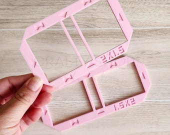 May include: A light pink plastic rectangular frame template with two open sections. The frame features embossed text reading 2X1.5 and 1.5X2, along with small triangular and M-shaped alignment markings along the edges of the frame structure.