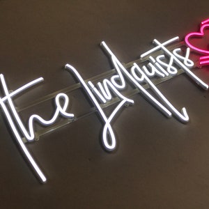 May include: Neon sign with the words "the lindquists" in white script. A pink heart with an arrow and other symbols is to the right. The sign is mounted on a clear plastic backing.