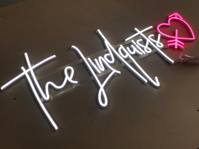 May include: Neon sign with the words "the lindquists" in white script. A pink heart with an arrow and other symbols is to the right. The sign is mounted on a clear plastic backing.