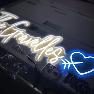 May include: A neon sign with the words "The Gravelles" in a cursive font, illuminated in white. A blue heart pierced by an arrow is on the right. The sign is on a light-colored surface.