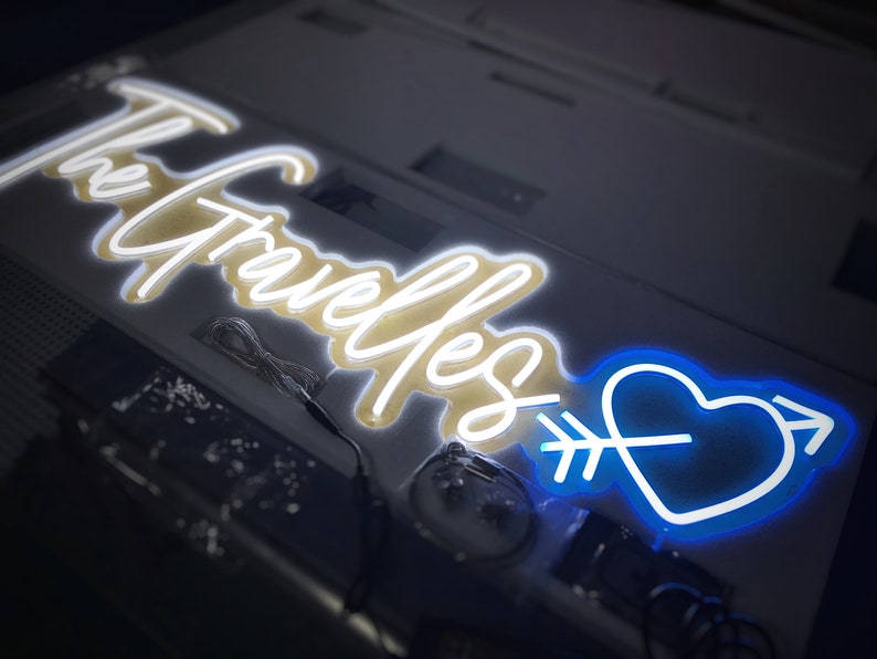 May include: A neon sign with the words "The Gravelles" in a cursive font, illuminated in white. A blue heart pierced by an arrow is on the right. The sign is on a light-colored surface.