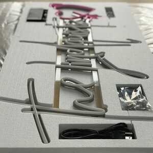 May include: A gray neon sign with the word "Dream" in a cursive font. The sign is in a white foam packaging with a power cord and small hardware. The word "Dream" is in a gray neon color, with a pink neon accent.