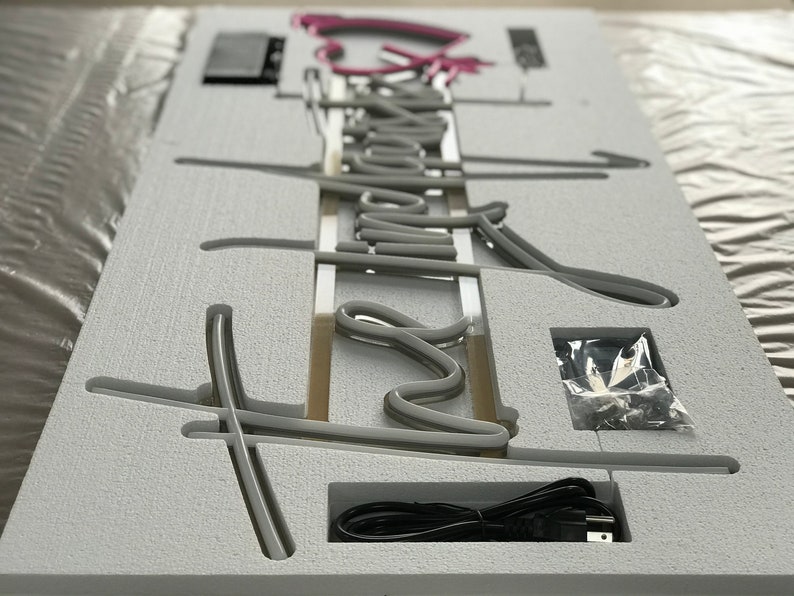 May include: A gray neon sign with the word "Dream" in a cursive font. The sign is in a white foam packaging with a power cord and small hardware. The word "Dream" is in a gray neon color, with a pink neon accent.