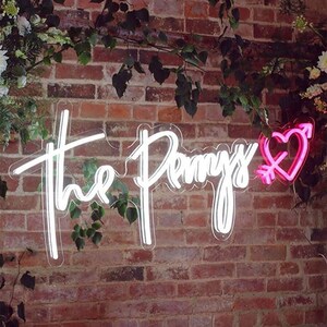 May include: A neon sign on a brick wall. The sign says "The Pennys" in a cursive font, with a pink heart pierced by an arrow. Green foliage surrounds the sign.