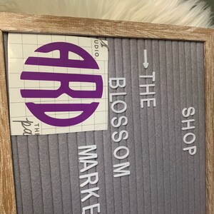 Op de afbeelding: Een paars monogram-letterontwerp op een witte rasterachtergrond met de tekst "The Blossom Market Shop" op een grijs letterbord.