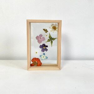 Op de afbeelding: Een houten frame met een heldere glazen inzet met geperste bloemen. De bloemen zijn in tinten roze, paars, blauw, oranje en wit.