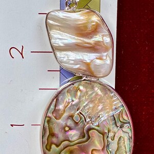 May include: A silver pendant featuring an iridescent abalone shell circular base and a smaller, irregular cream-colored mother-of-pearl top section. A ruler placed behind the pendant shows a total length of approximately 2.5 inches.