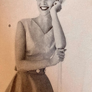 May include: A vintage black and white photograph of a woman wearing a V-neck top and a flared skirt. The top has a sleeveless design, and the skirt is buttoned at the waist. The woman is smiling and wearing a headscarf.