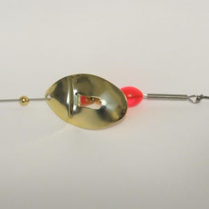 May include: A gold fishing lure with a red bead and a single hook. The lure has a unique shape with a hole in the center.