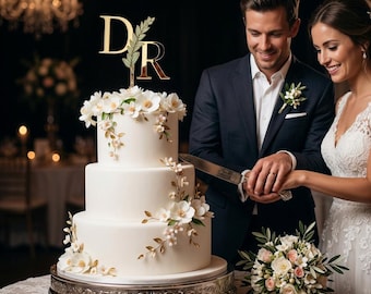 May include: A three-tiered white wedding cake decorated with white flowers and gold-toned leaves. A gold cake topper features the letters D and R separated by a green leafy branch. The cake sits on a silver pedestal stand on a lace-covered table.