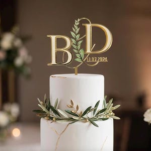 Può includere: Un topper per torta in acrilico color oro con le lettere B e D in carattere serif, collegate da un design a ramo di foglie verdi. La data 11.07.2024 è incisa alla base. Il topper è posizionato su una torta bianca con accenti dorati.