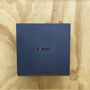 Puede incluir: Un dispositivo rectangular gris oscuro con la palabra "BOBCAT" impresa en rojo en la parte delantera.